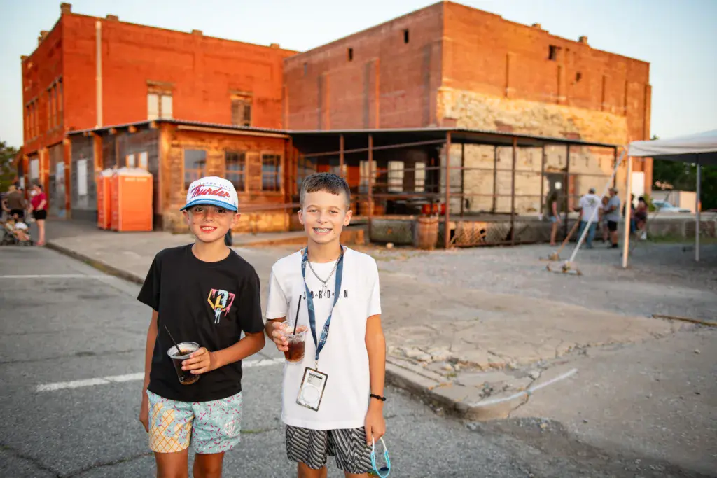 Two young festival attendees smile at the camera in front of rustic red-brick festival venue buildings.