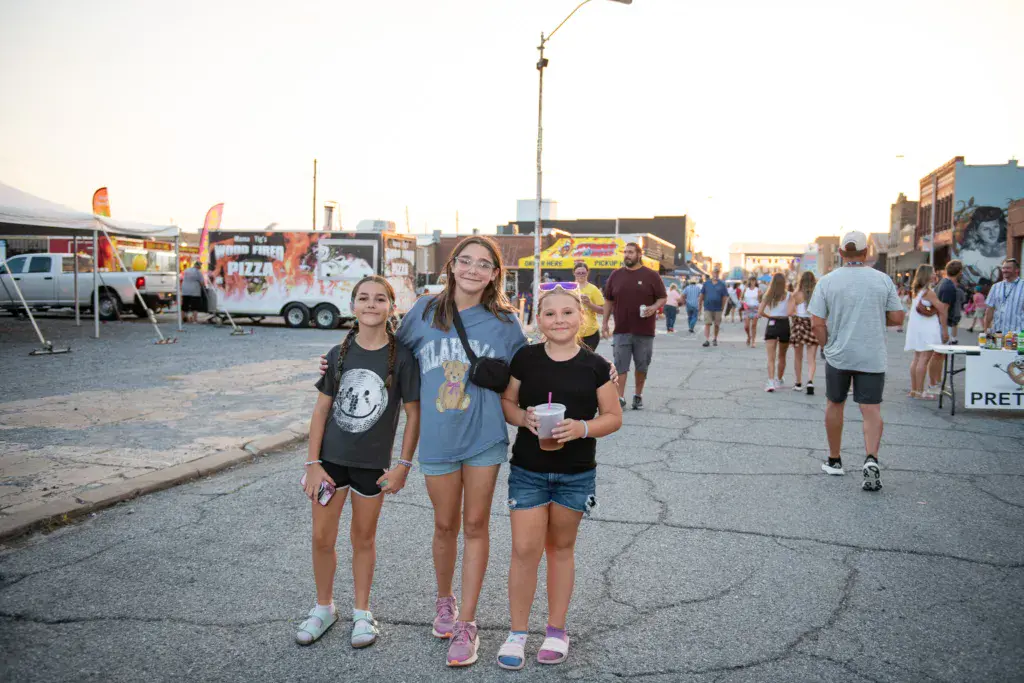 Three young attendees stand together in a festival grounds with food vendors and crowds visible behind them.