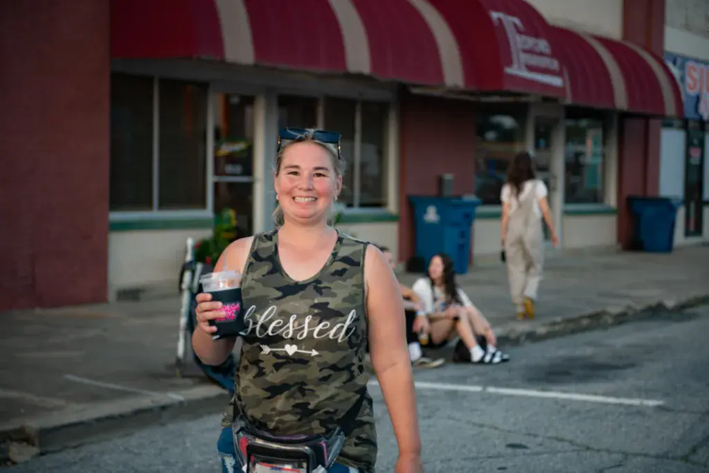 A smiling attendee in a camouflage tank top holds a drink at the Dancing Rabbit Music Festival venue.