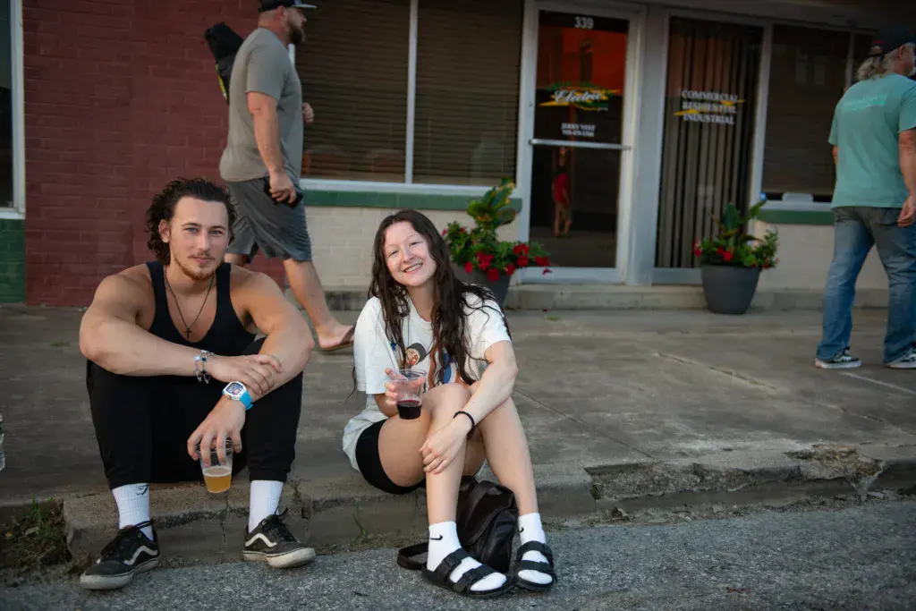 Two festival attendees sit together on pavement holding drinks in front of a building.