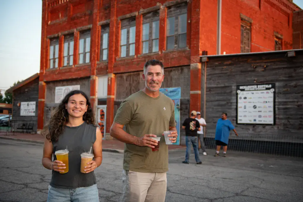 Two people holding drinks smile at an outdoor festival venue with a red brick building and other attendees in the background.