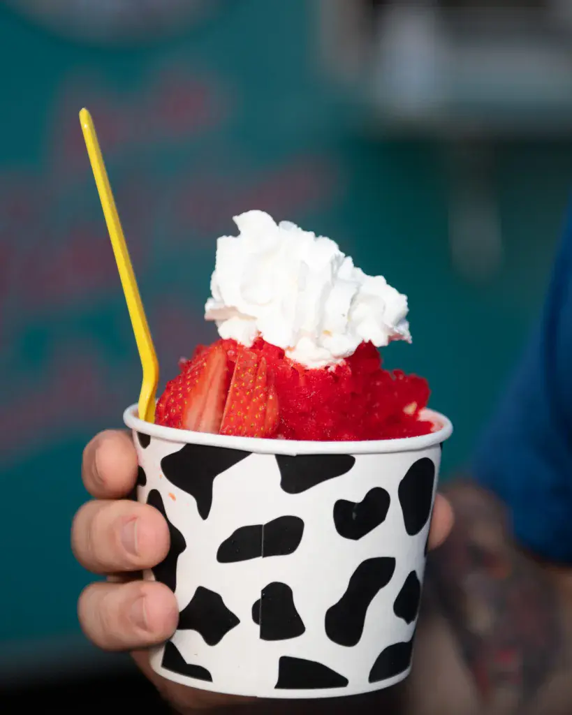 A hand holds a cow-print cup filled with red fruit, whipped cream, and a yellow straw at the festival.