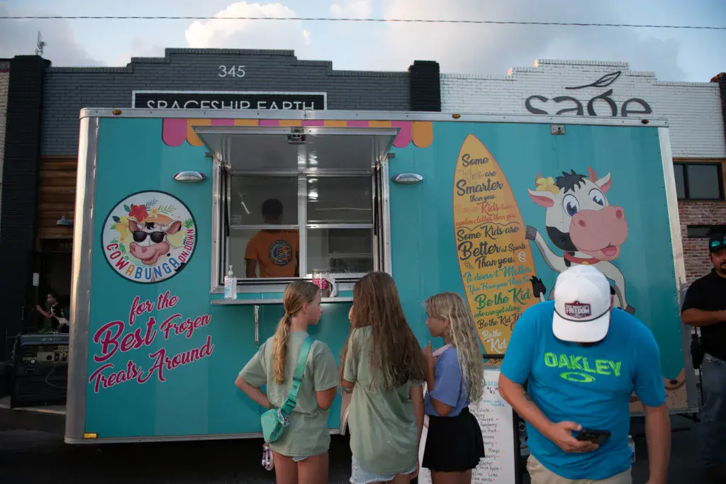 Festival attendees order frozen treats from a colorful turquoise food vendor truck with playful graphics and signage.