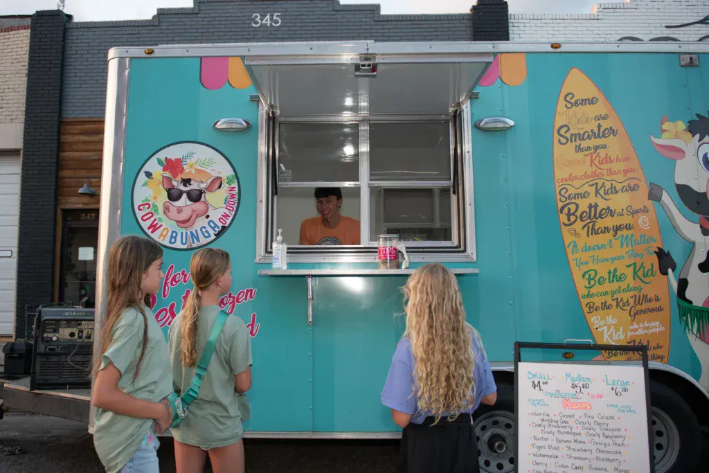 Three young people wait at a colorful turquoise food truck window at an outdoor festival.