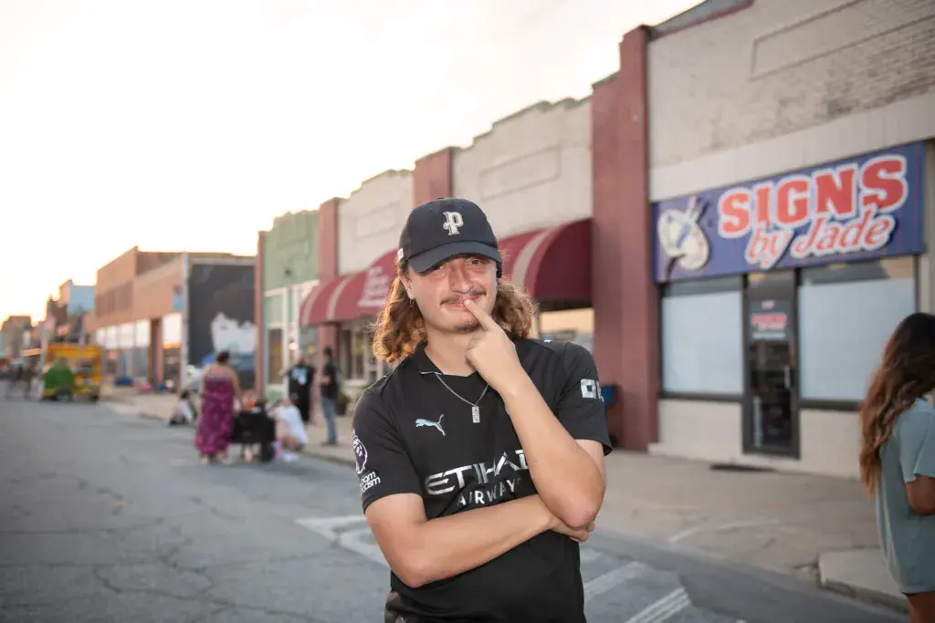 A person in black attire and cap poses thoughtfully in front of storefronts at the festival venue.