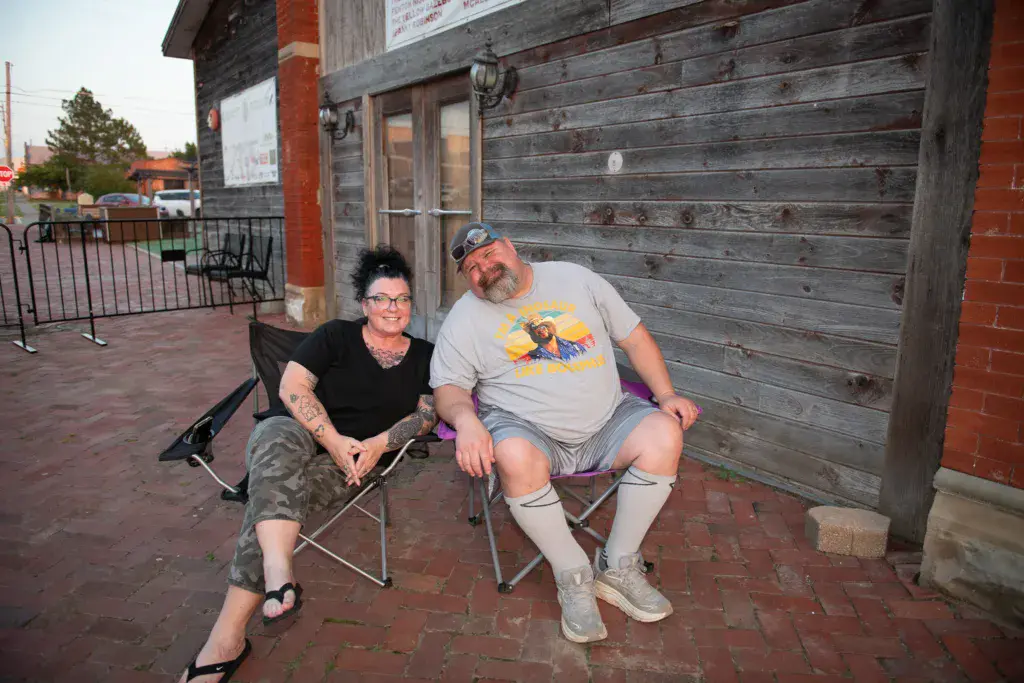 Two people sit in folding chairs outside a wooden building at the festival venue.