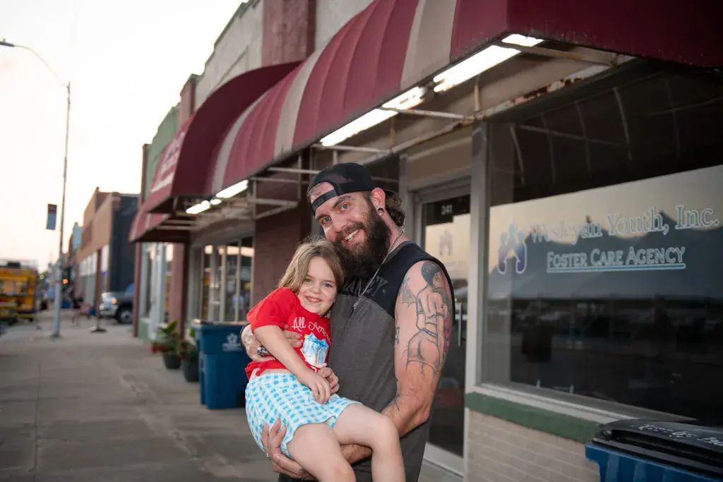 A bearded man wearing a black cap and sleeveless shirt holds a young child in a red shirt on an outdoor downtown street.