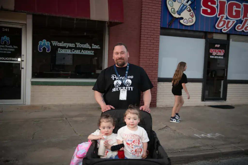 A festival security staff member poses with two young children in a stroller outside a brick storefront.