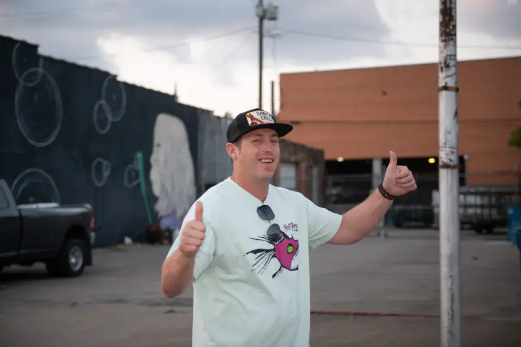 A man in a light-colored graphic t-shirt and black cap gives double thumbs up while smiling at an outdoor venue.