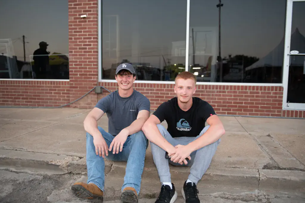 Two smiling men crouching on a concrete patio in front of a brick building at dusk.