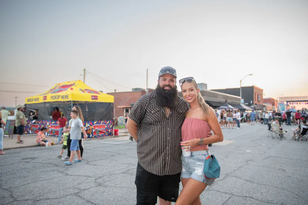 Two people smile at the camera in the festival grounds with a yellow vendor tent and crowd visible behind them.