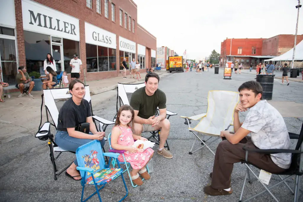 A family of five sits together in camping chairs on a street at an outdoor festival, with historic brick buildings in the background.