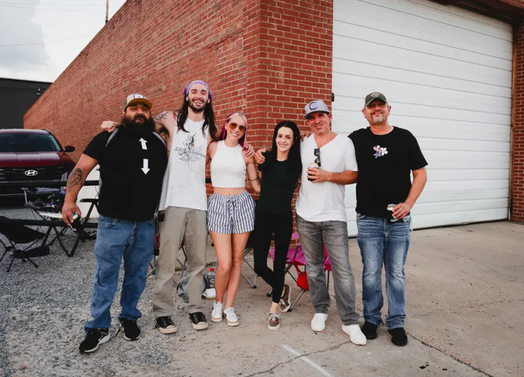 Six festival attendees stand together smiling in front of a brick building and garage door.