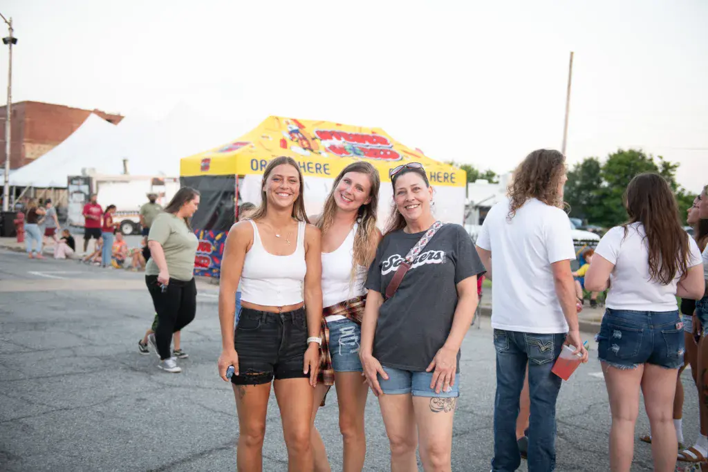 Three smiling women pose together at an outdoor music festival with crowds and vendor tents visible behind them.