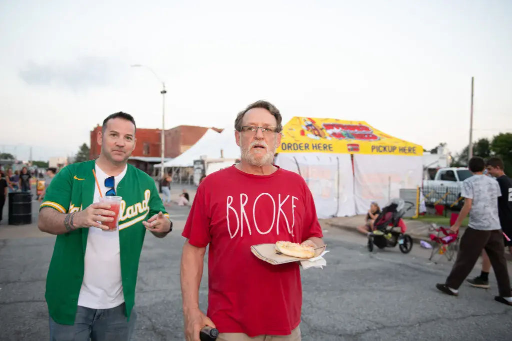 Two attendees walk through the festival grounds holding food and drinks near a colorful vendor tent.