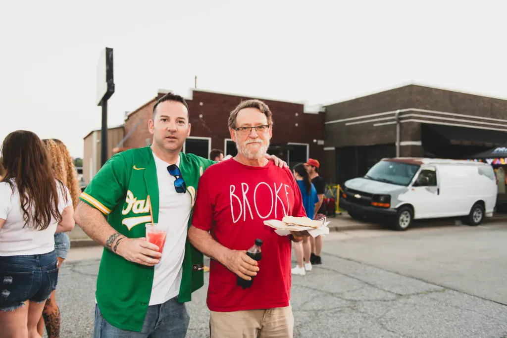 Two men holding food and drinks stand in a parking lot during the festival with buildings and a white van in the background.