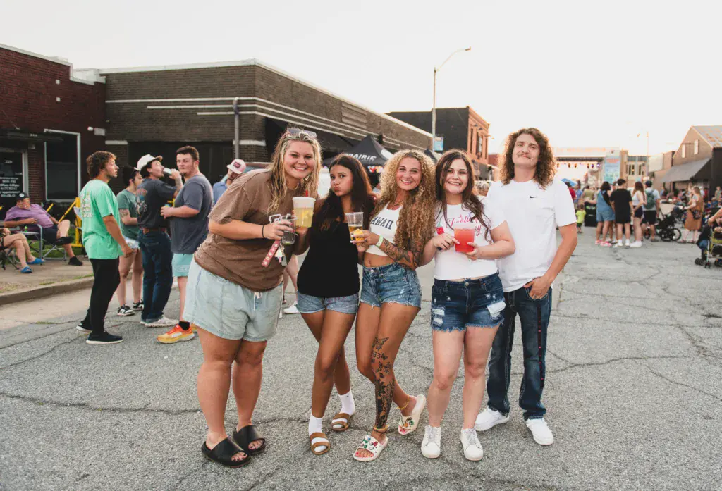 A group of six young people pose together in the street at an outdoor music festival venue with brick buildings and festival crowd in the background.