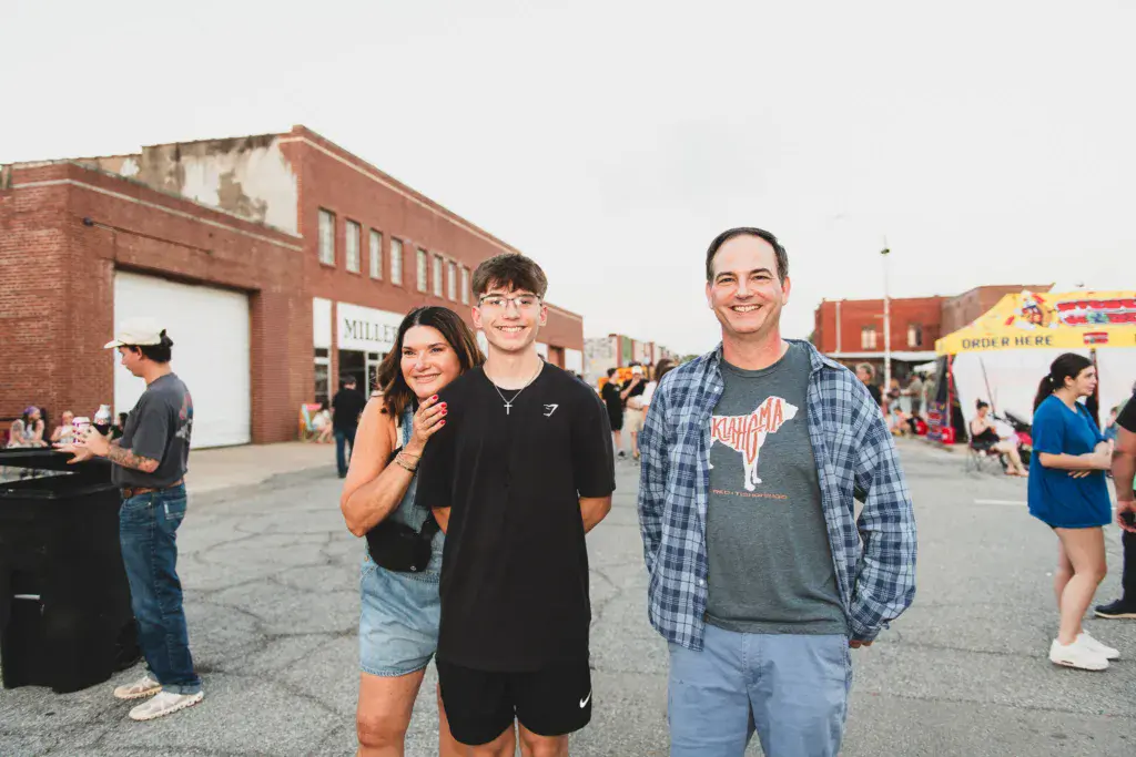 Three smiling attendees pose in front of a brick building at an outdoor music festival venue.