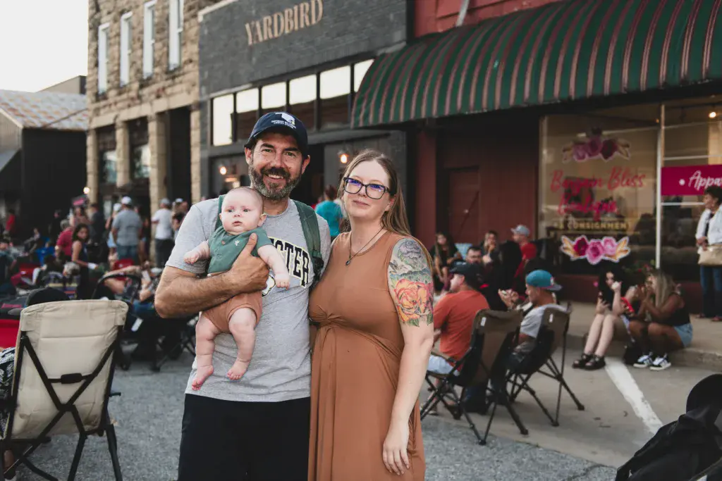 A family of three stands together on a street in front of a brick building at an outdoor festival.