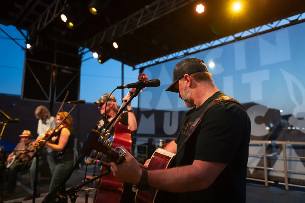 A band performs on an outdoor stage at dusk with stage lighting and a large screen backdrop.