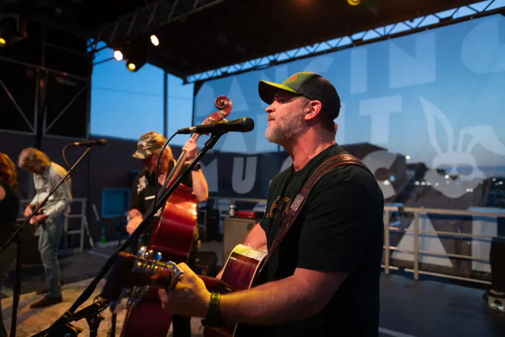 A performer wearing a black cap and shirt sings into a microphone while playing an acoustic guitar on stage at dusk.