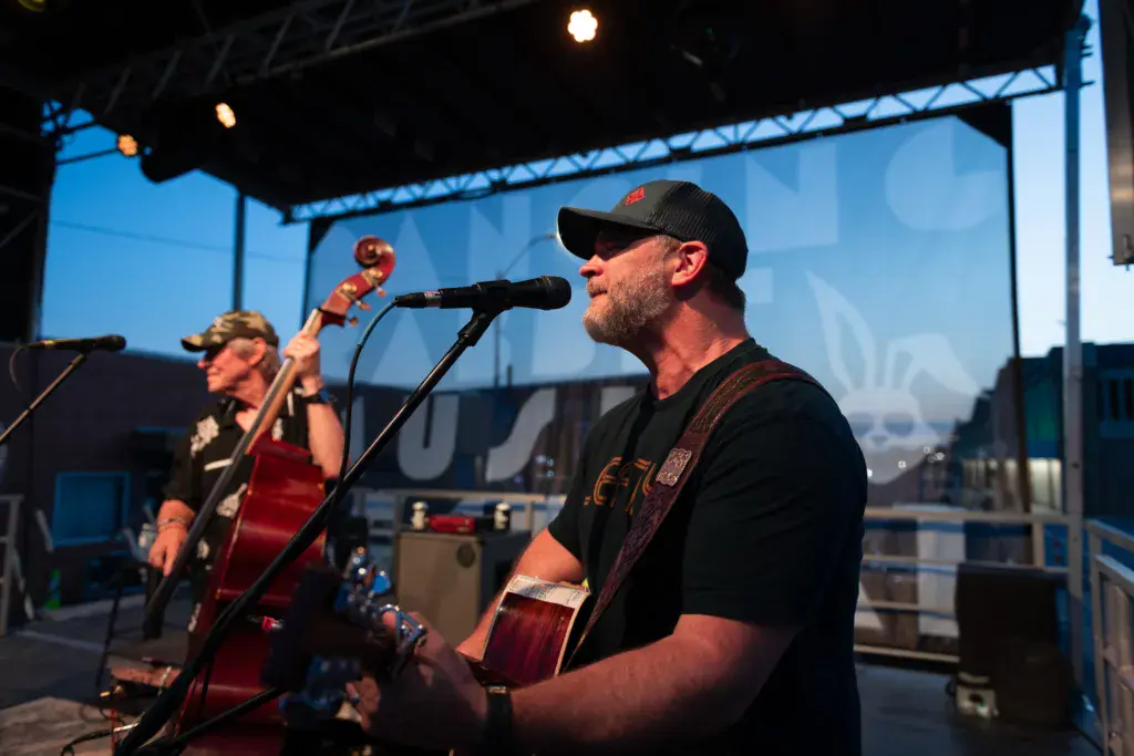 A musician performs at an outdoor festival stage, singing into a microphone while playing an acoustic guitar.
