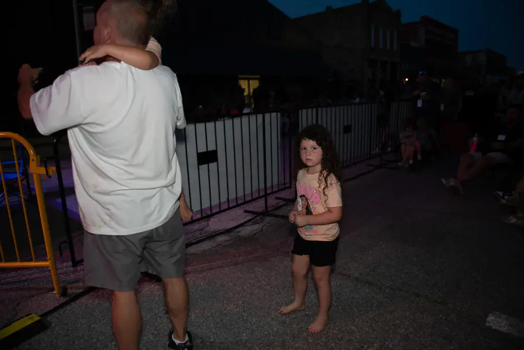A child and adult walk through a festival venue at dusk near metal barriers and buildings.
