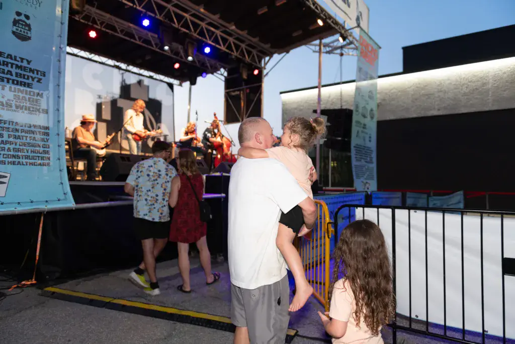 A man holds a child while watching a live band perform on an outdoor festival stage.