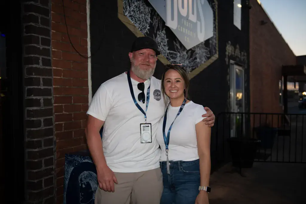 Two festival staff members wearing lanyards pose together outside a brick venue building at dusk.