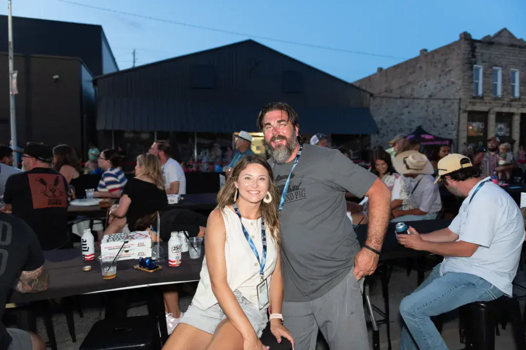 Two people smile at the camera during a daytime festival gathering with crowds and buildings in the background.