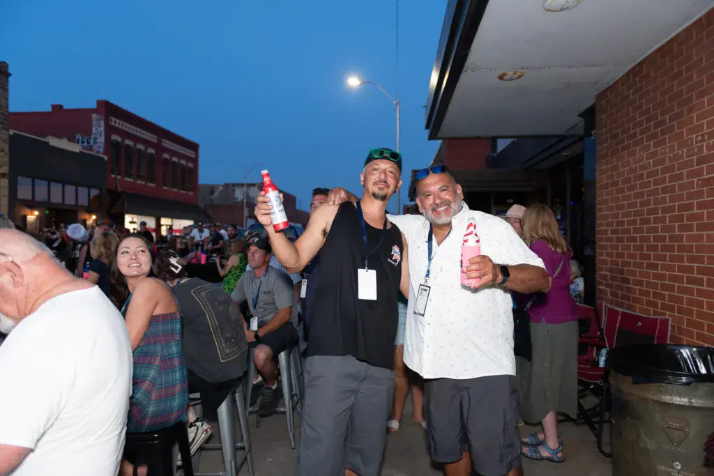 Two men wearing festival badges celebrate together outdoors at dusk, holding beverages with a crowd and brick buildings in the background.