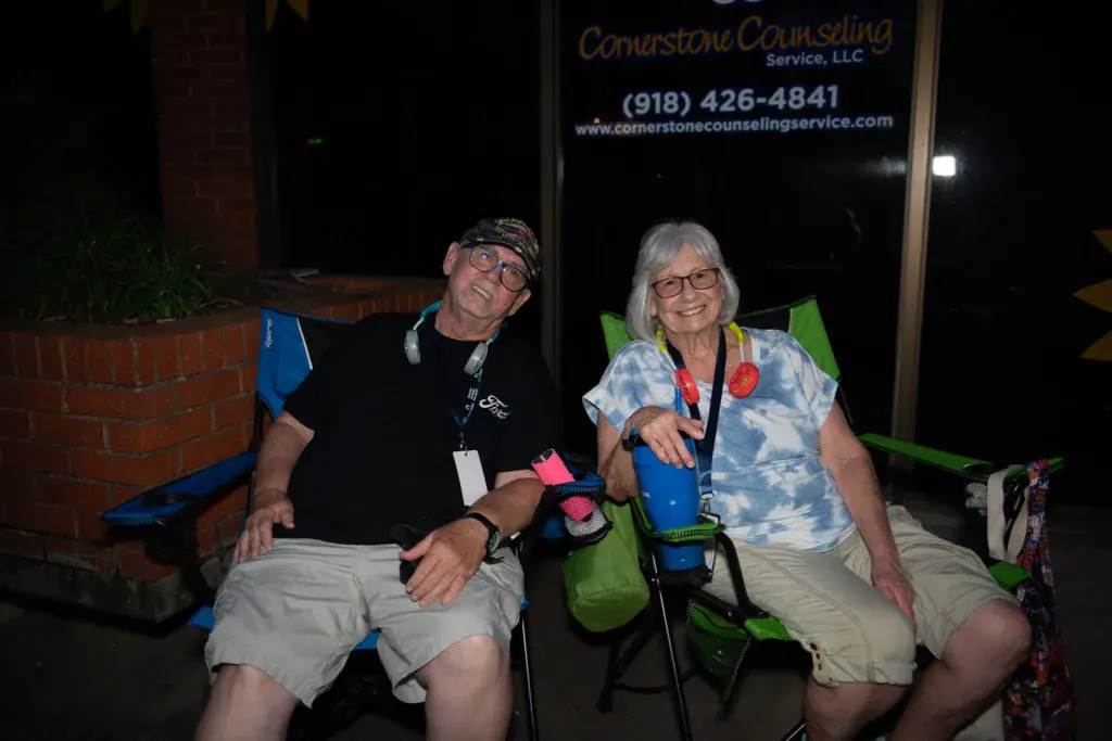 Two festival attendees sit together in lawn chairs at night, wearing event badges and smiling at the camera.