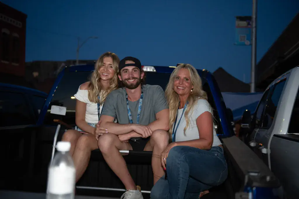 Three people wearing festival lanyards sit together in the bed of a pickup truck at dusk.