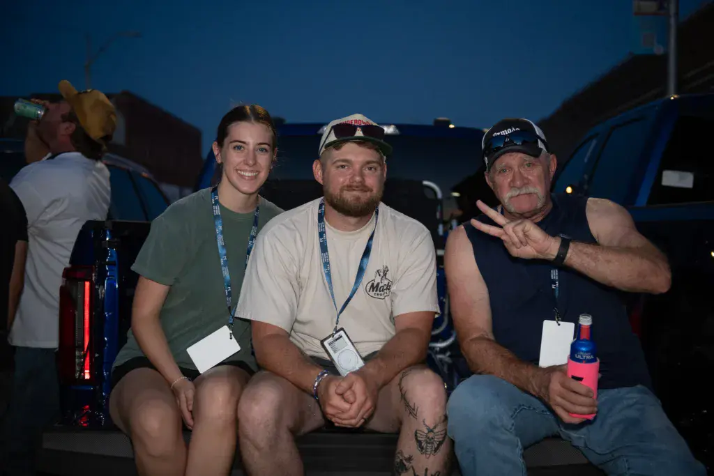 Three people wearing festival laminates sit together at dusk with festival structures visible in the background.