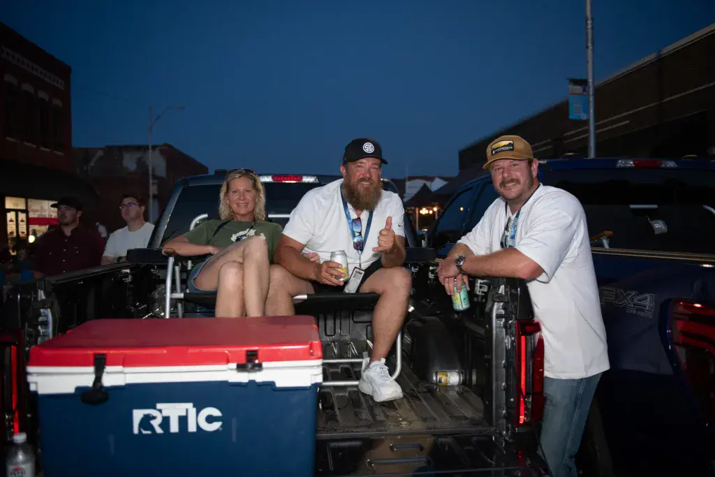 Three people sit in the bed of a pickup truck with an RTIC cooler at dusk during the festival.
