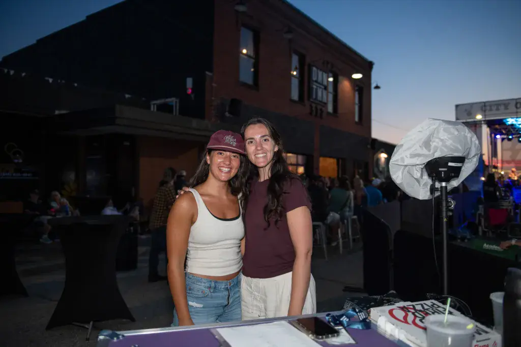 Two people smile at the camera behind a festival booth with equipment and signage during evening at Dancing Rabbit Music Festival.