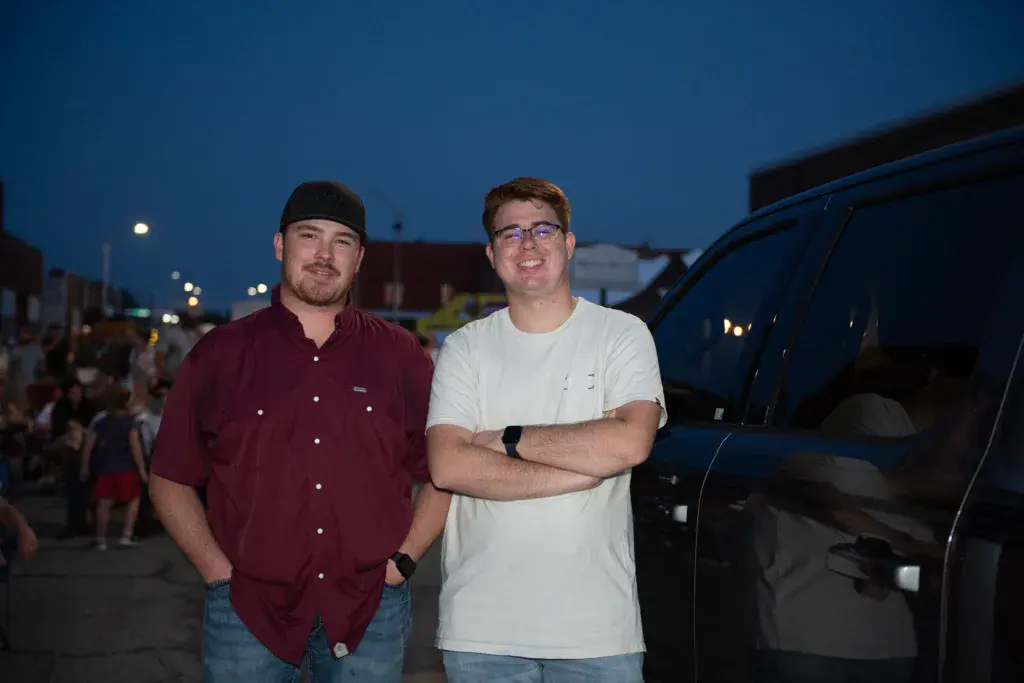 Two people stand together in a parking lot at dusk during the festival, with vehicles and crowds visible behind them.
