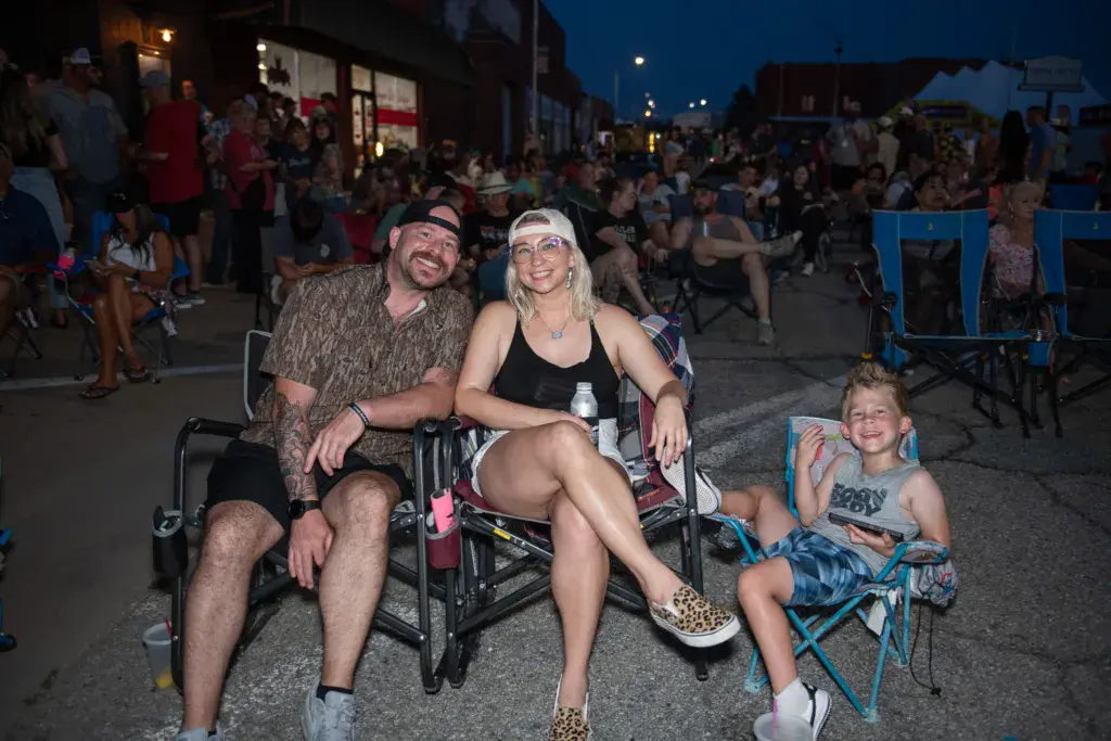 A family of three smiles at the camera while seated in camping chairs at the Dancing Rabbit Music Festival during evening.