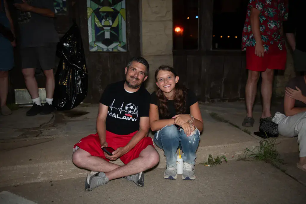 Two people smile at the camera while crouching together at a nighttime outdoor event.
