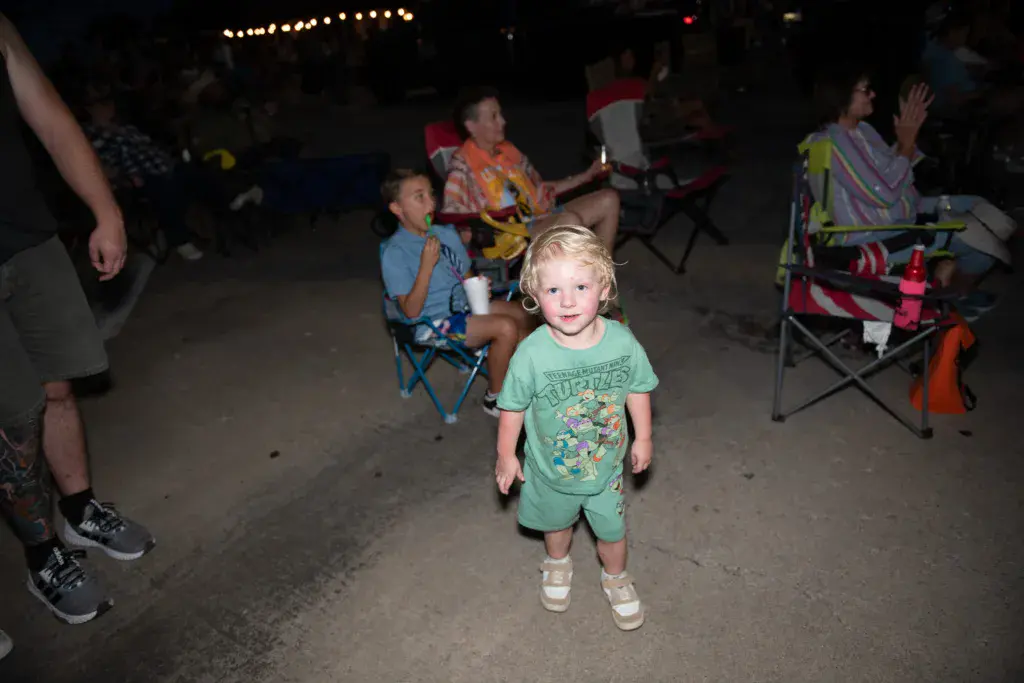 A young child in a green shirt walks through a nighttime festival crowd with camping chairs and attendees around them.