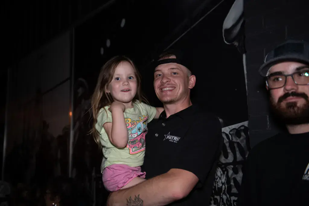 A man wearing a black polo shirt holds a young child at an outdoor festival at night, with another man visible to the right.