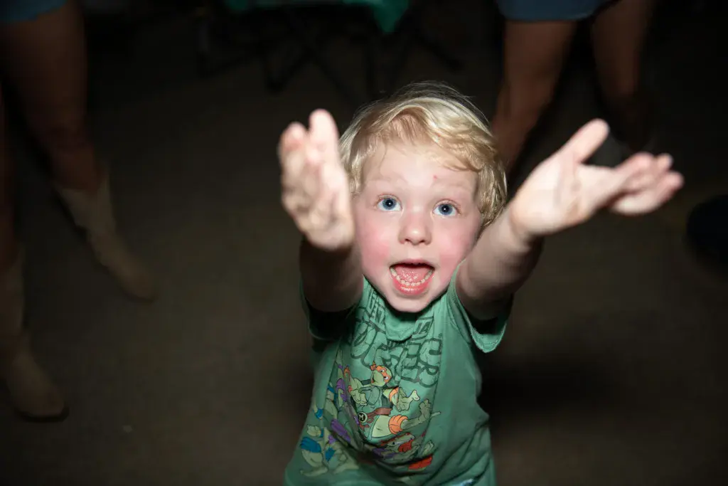 Young child with blonde hair in green shirt raising arms excitedly at night event.