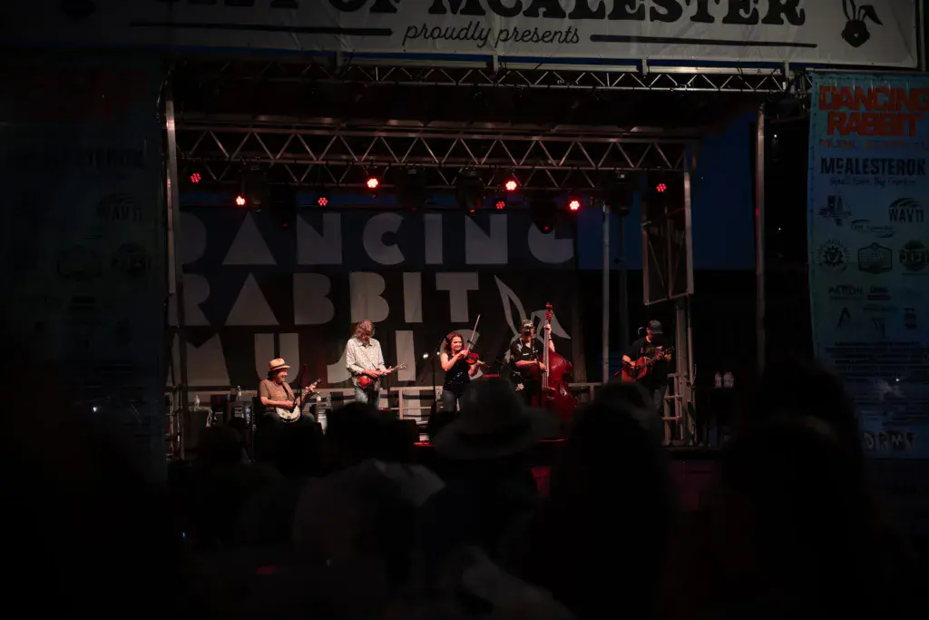 A band performs on an illuminated stage at night while audience members watch from below.