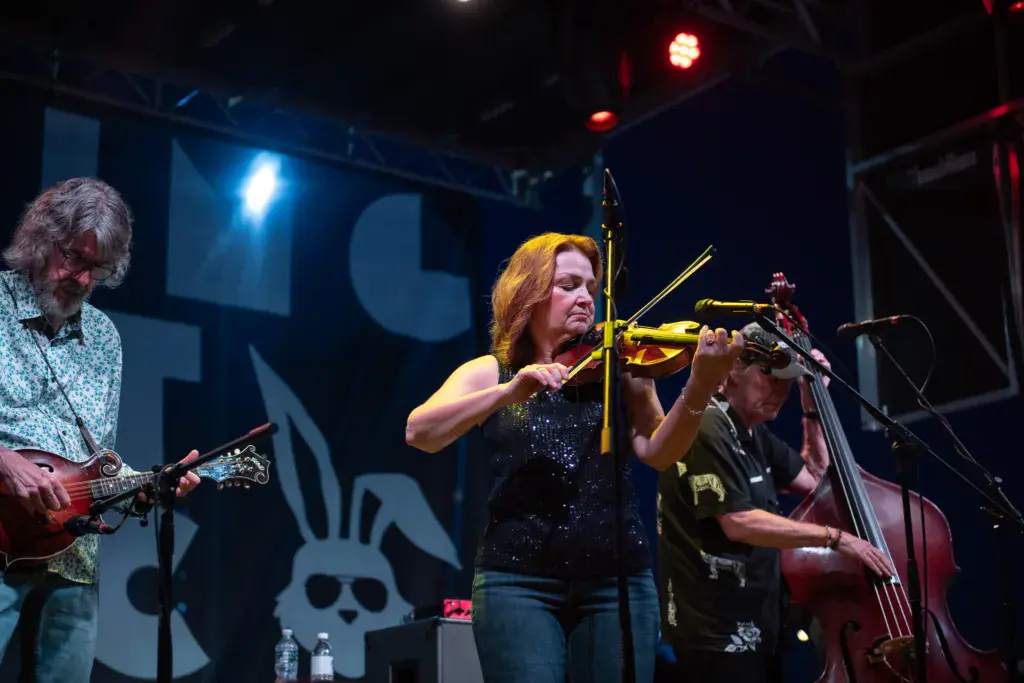 A band performs on stage with a violinist in a sparkly top, guitarist, and bassist under blue and red stage lighting.