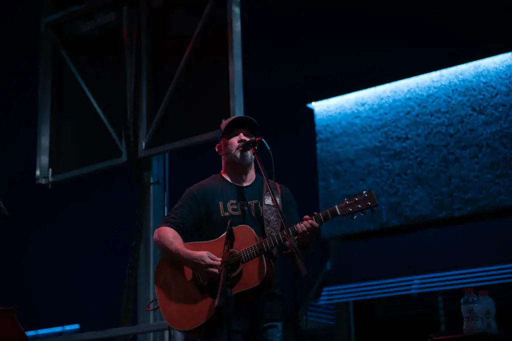A bearded musician wearing a cap and dark shirt plays an acoustic guitar at night on an illuminated stage.
