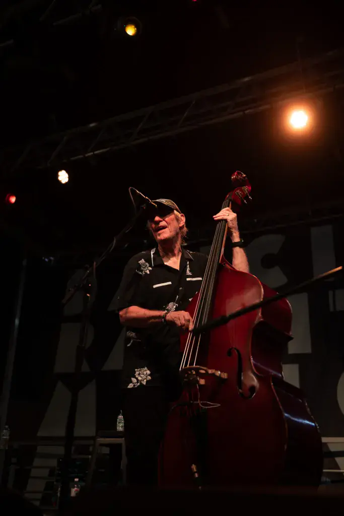A musician wearing a black shirt and cap plays a red cello on stage under warm stage lighting.
