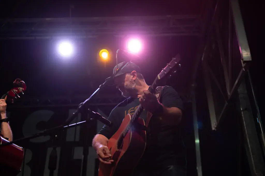 A performer plays an acoustic guitar at night on stage under bright pink and white lights.
