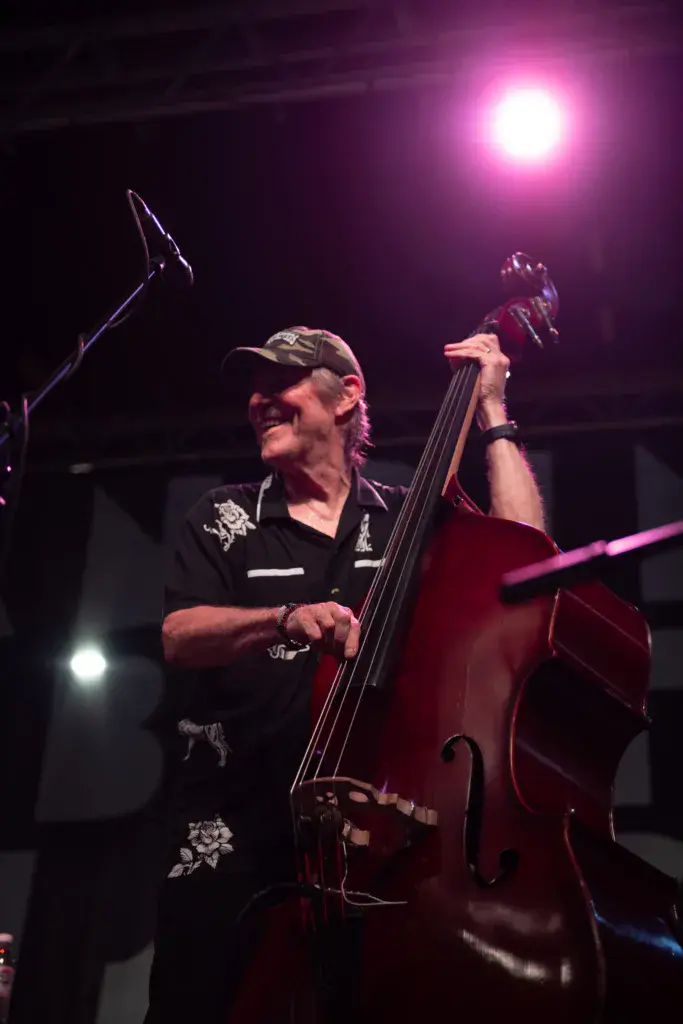 A bass player performs on stage wearing a black shirt and cap, holding a red cello bass under stage lights.