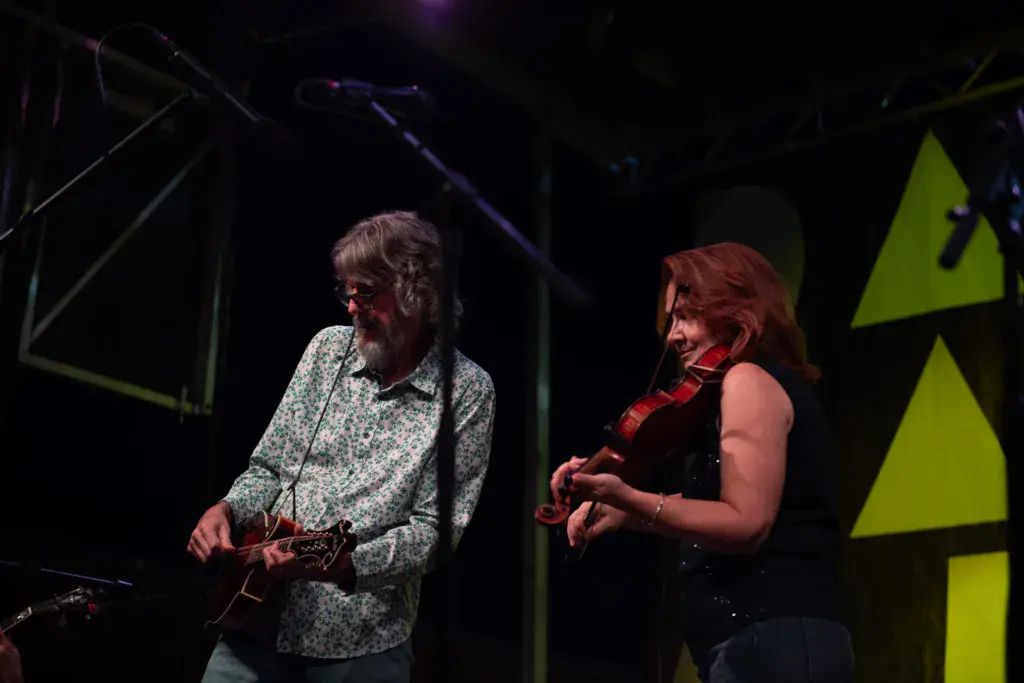 Two musicians perform on stage under stage lighting with geometric backdrop elements behind them.