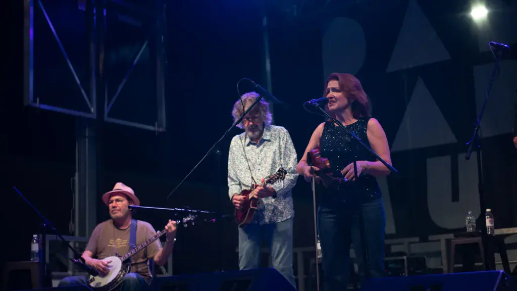 Three musicians perform on stage during an evening concert at the festival, with stage lighting and geometric backdrop structures behind them.
